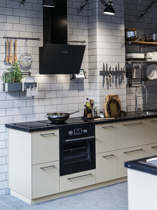 A modern kitchen with HAVSTORP beige doors and black appliances in a white tiled room.