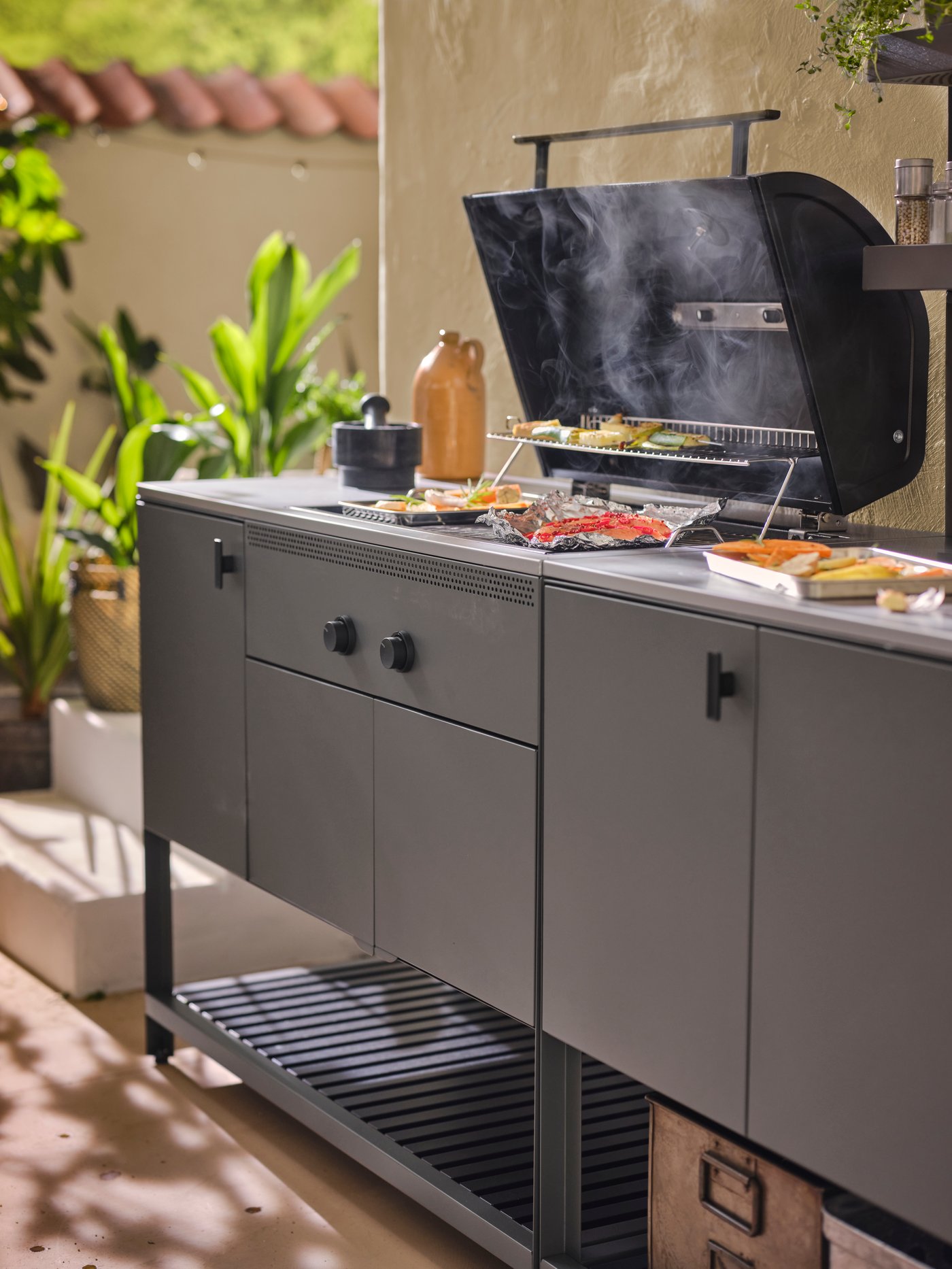 A BÅTSKÄR charcoal barbecue with some food on the grill and some vegetables on the worktop in a GRILLTIDER serving tray.