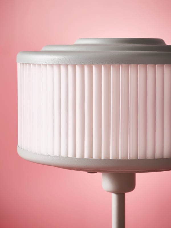 The pleated white lampshade of a grey-green REMTÅNG desk lamp against a soft pink backdrop.