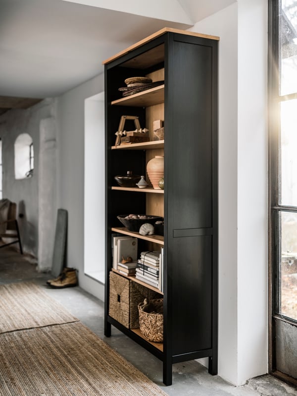 Valoisassa tilassa mustanruskea HEMNES-kirjahylly, jonka hyllyillä on koriste-esineitä, kirjoja ja koreja.