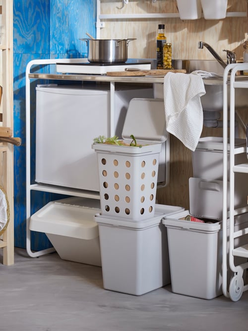 A mini-kitchen setup with a waste sorting bin with lid, two bins with lids, a bedspread, and a pot with lid.