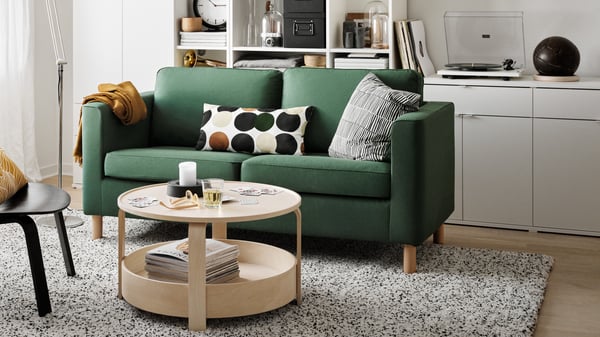 Modernes Wohnzimmer mit grünem Zweisitzer-Sofa, gemusterten Kissen und rundem Couchtisch, im Hintergrund Regalwand mit Plattenspieler