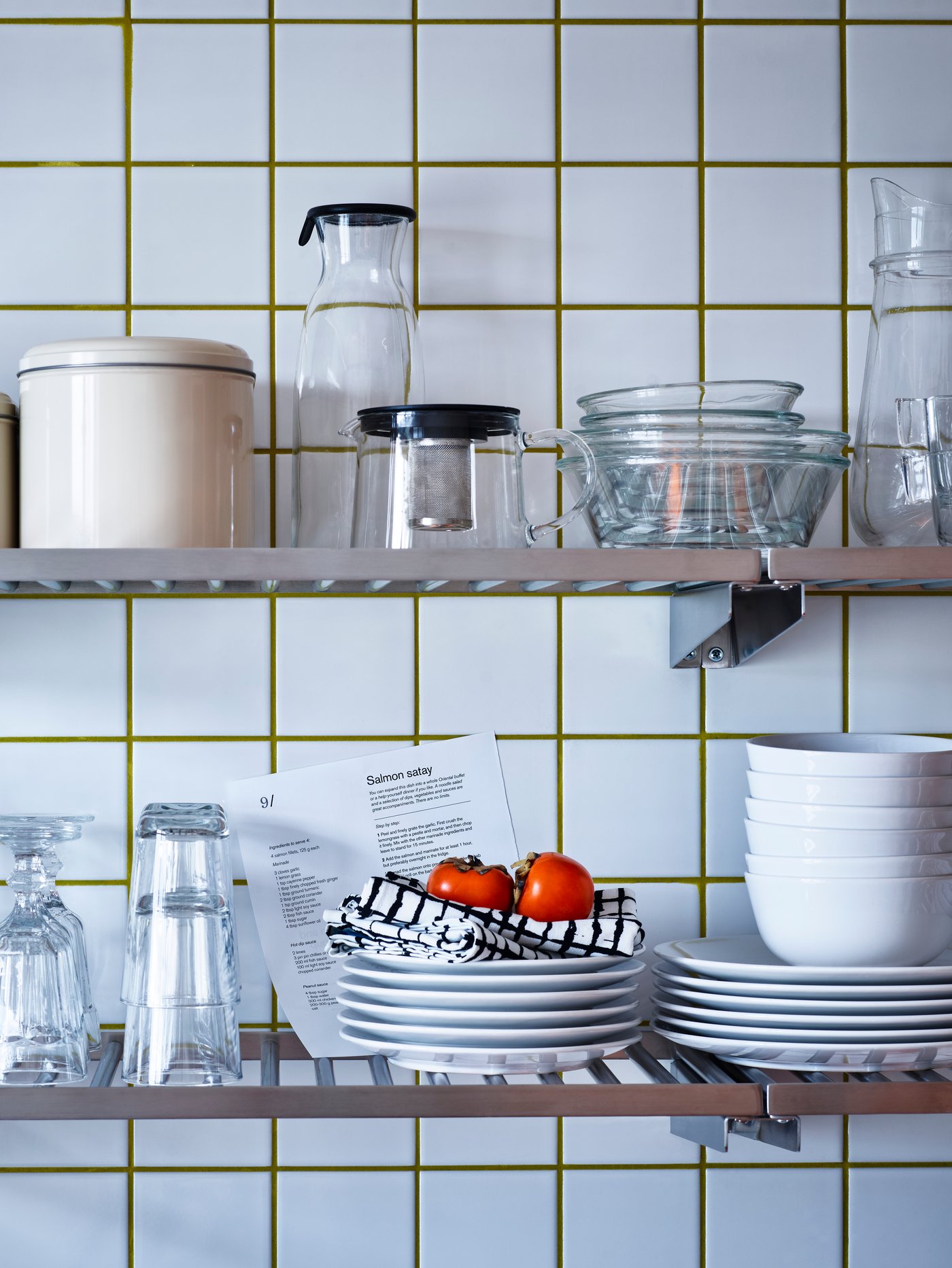 Vista de una cocina que se centra en una pared de azulejos con dos estantes con vasos, cuencos, platos, una tetera y una jarra con tapa.