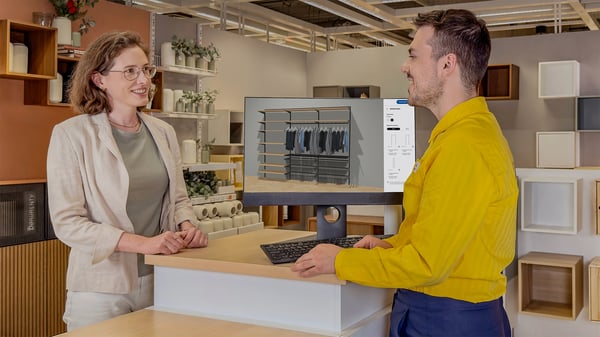 Konsultant IKEA stojący w żółtej koszulce przy stoisku planowania rozwiązań do przechowywania. Przy stoisku stoi również klientka, która omawia z konsultantem projekt rozwiązania na ekranie komputera.