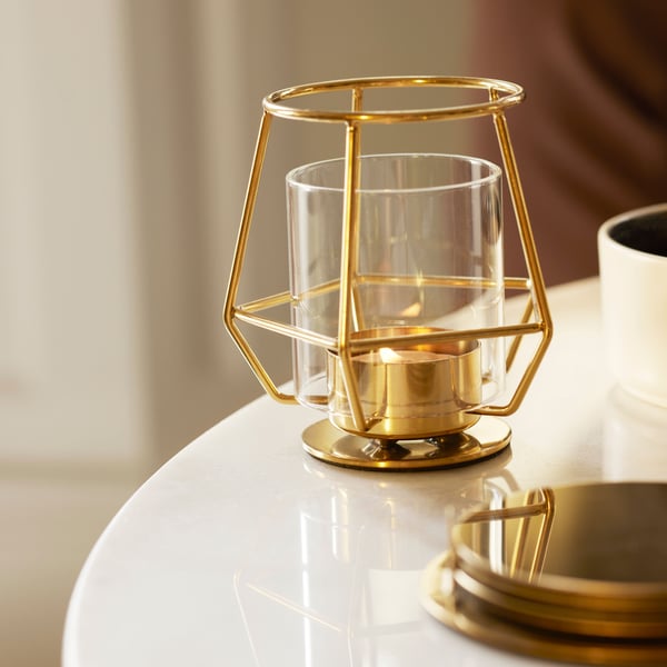 A close-up of a table top with an airy, geometric-shaped PÄRLBAND tealight holder in gold-coloured stainless steel.