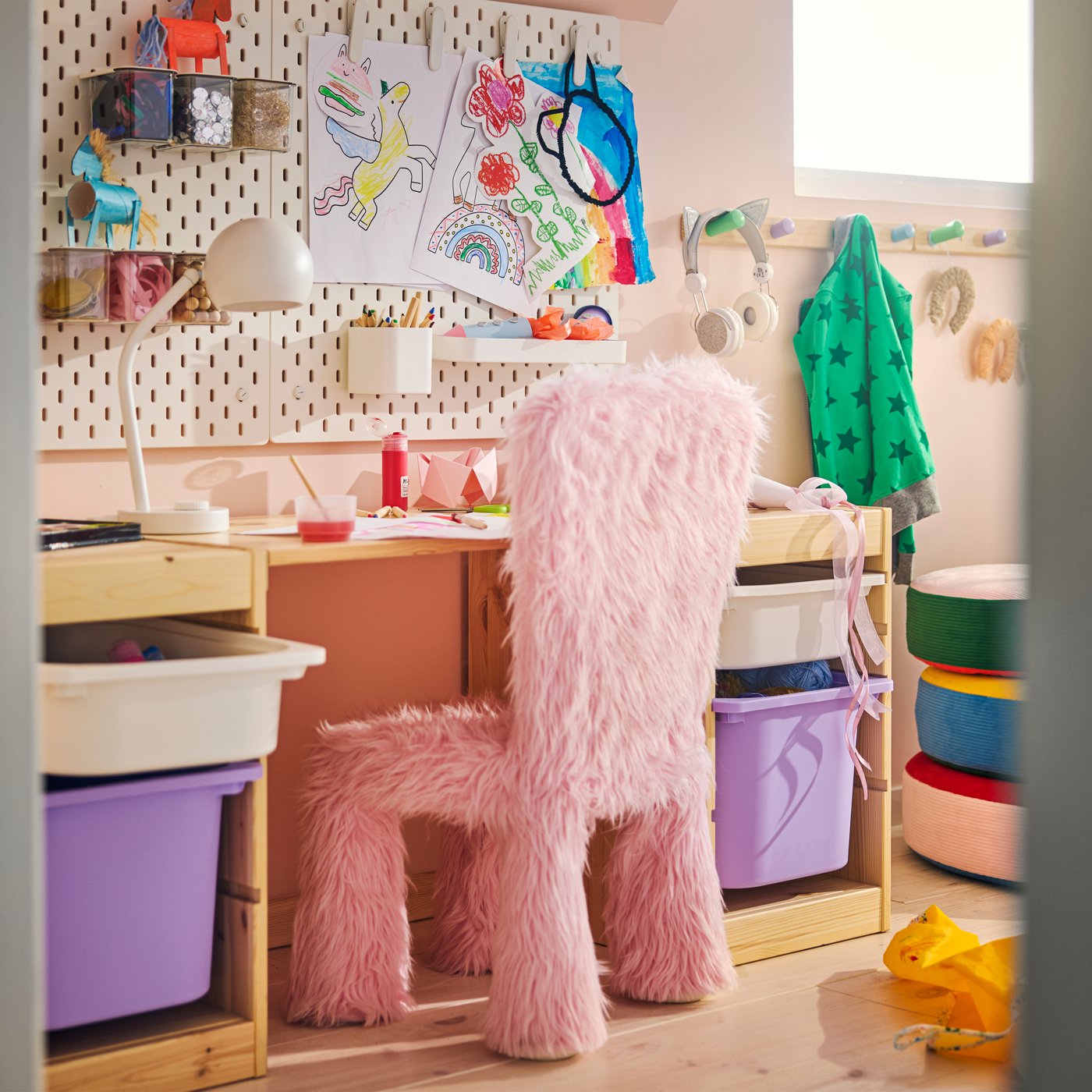 ピンクのフェイクファーGREJSIMOJS 子ども用チェアカバーがある子ども部屋で、ペグボードの壁のそばに机と収納ユニットが並んでいる。