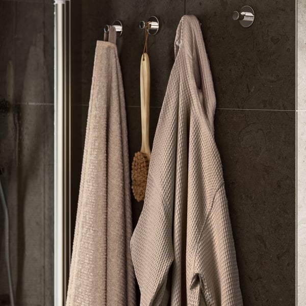 A beige, waffle-textured BJÄLVEN bathrobe hangs on a hook on the wall beside a beige VALLASÅN towel and a wooden body brush.