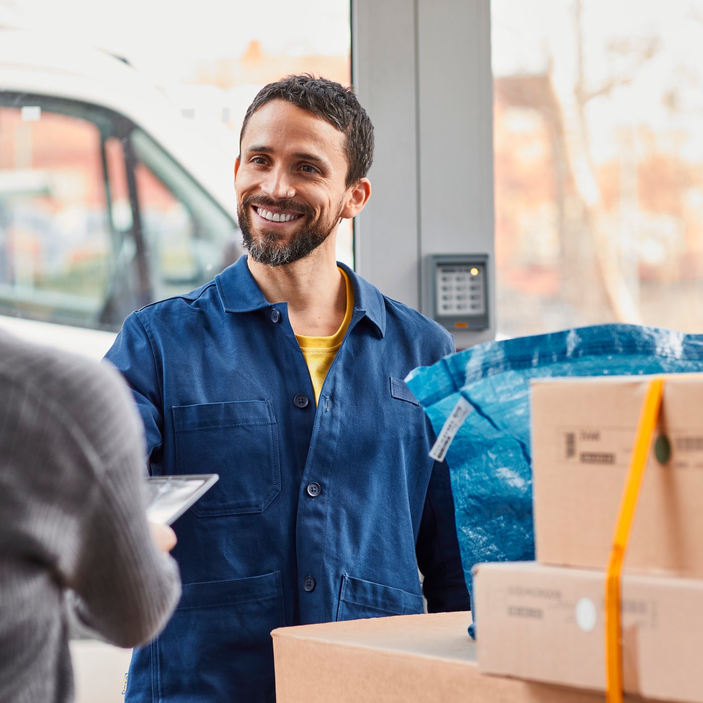 En medarbeider i en blå skjorte leverer noen pakker til en kvinne i forgrunnen med en grå genser.