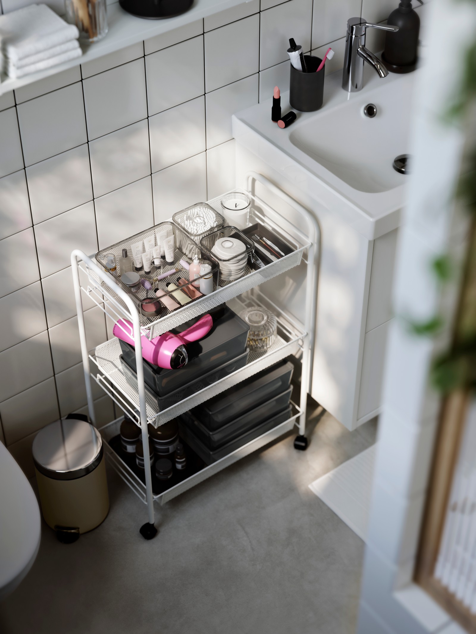 A white HORNAVAN trolley with three shelves filled with bath accessories neatly organised. Shown in a modern bathroom.