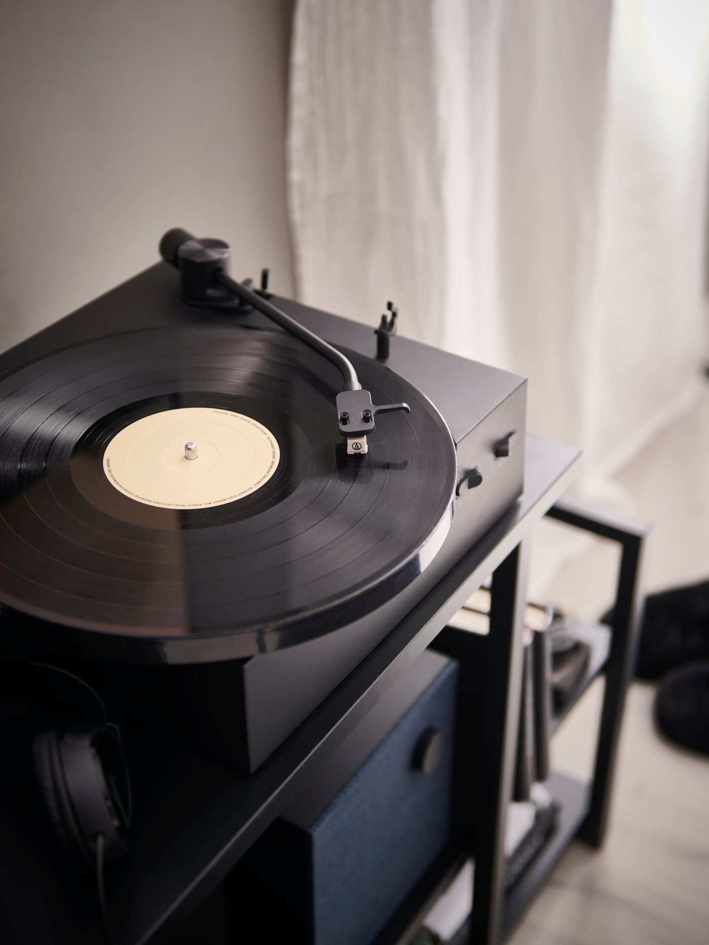 Un giradischi OBEGRÄNSAD sopra uno scaffale nero accanto a una finestra con una tenda bianca.