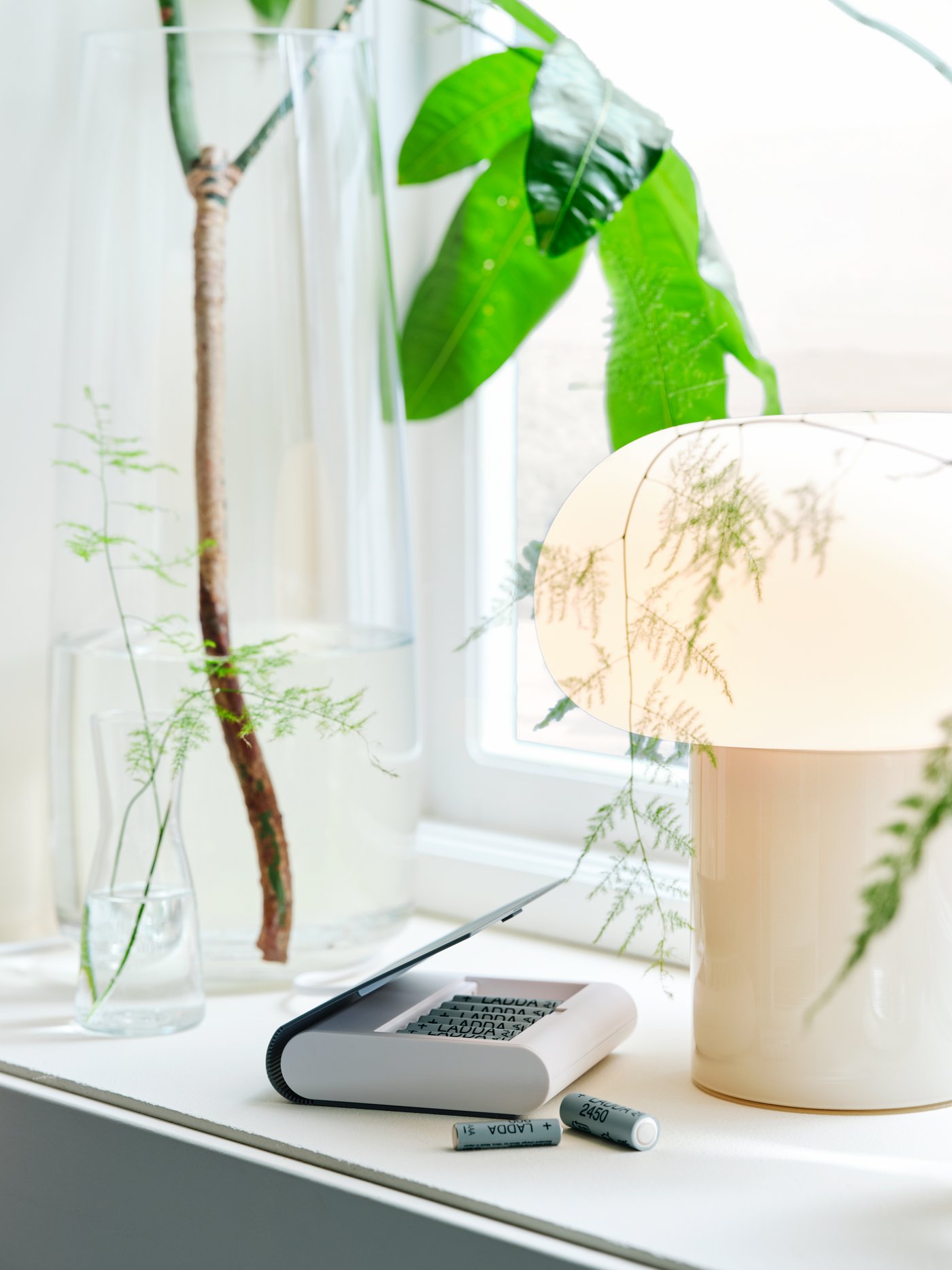 A DEJSA table lamp sits on a windowsill with a TJUGO battery charger with storage and two LADDA rechargeable batteries.