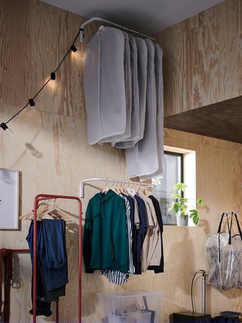 Un espace de rangement avec un portant, des sacs de rangement et deux tringles à vêtements accrochées au mur avec des habits sur des cintres et des housses.