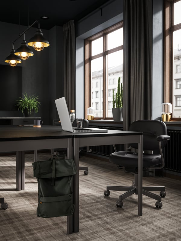 مكاتب IDÅSEN لون أسود/رمادي داكن وكرسي MULLFJÄLLET، وحقيبة ظهر باللون الأخضر الزيتوني في المقدمة ونوافذ في الخلفية.