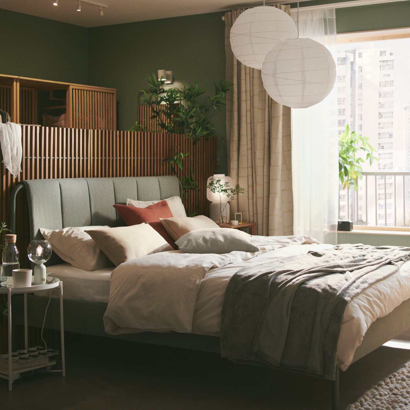 Una struttura letto imbottita TÄLLÅSEN grigio-verde con dietro dei paraventi, in una camera da letto verde con finestre ampie e un'atmosfera serena.