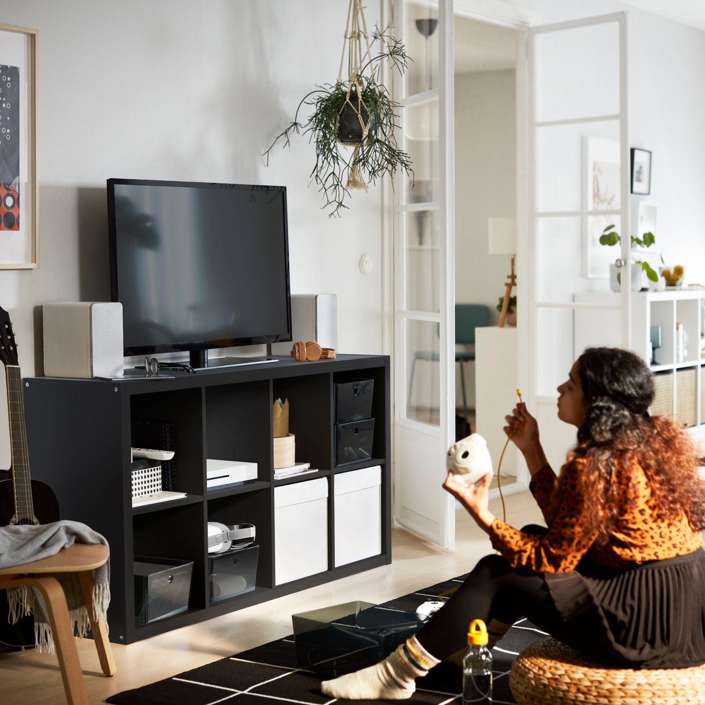 Person sitting on the floor in a bright living room, connecting a device to a TV on a black storage unit. Hanging plant, guitar, and organized shelves complete the modern space.
