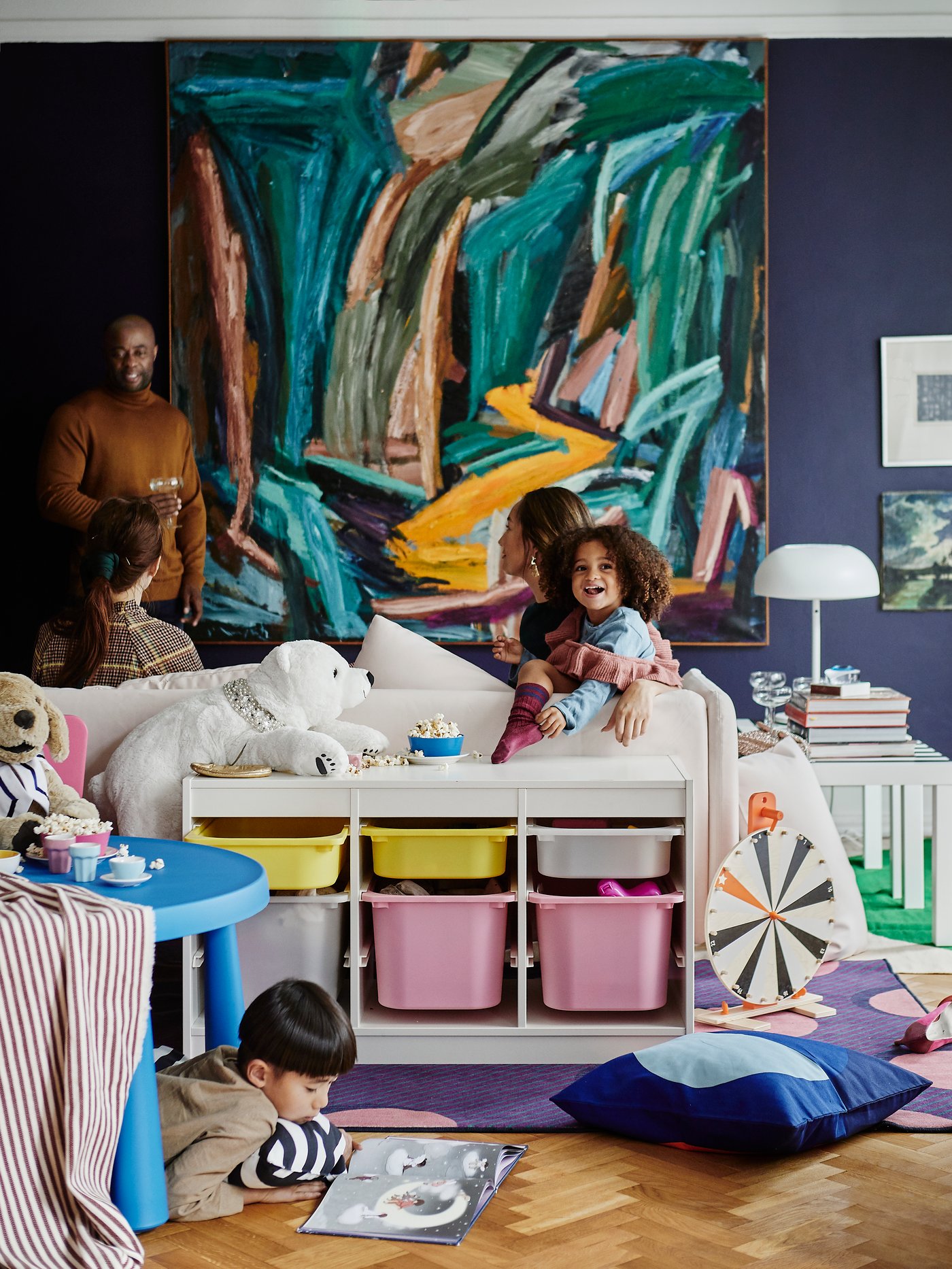 Et barn sidder i en sofa bag barnet sidder en kvinde og til højre står en mand på gulvet foran sofaen og en opbevaringsreol ligger en dreng og tegner og der er et stort maleri bag sofaen i det rodede rum.
