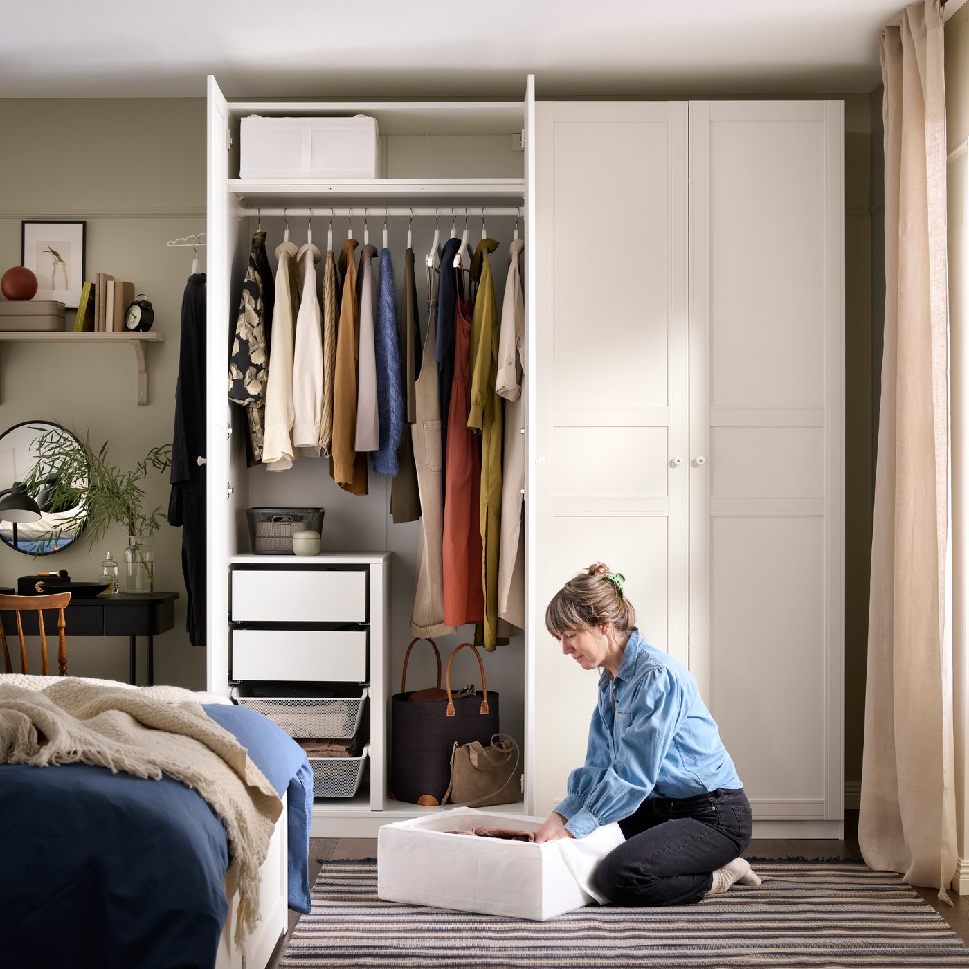 A bedroom with an open white PAX wardrobe showing clothes hanging, and a woman sitting on the floor organising a SKUBB box.