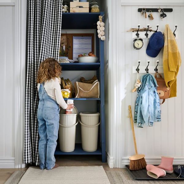 Dívka odhrnuje závěs, aby se dostala ke spíži s úložným dílem IVAR a košům DAMMÄNG sloužícím k recyklaci.