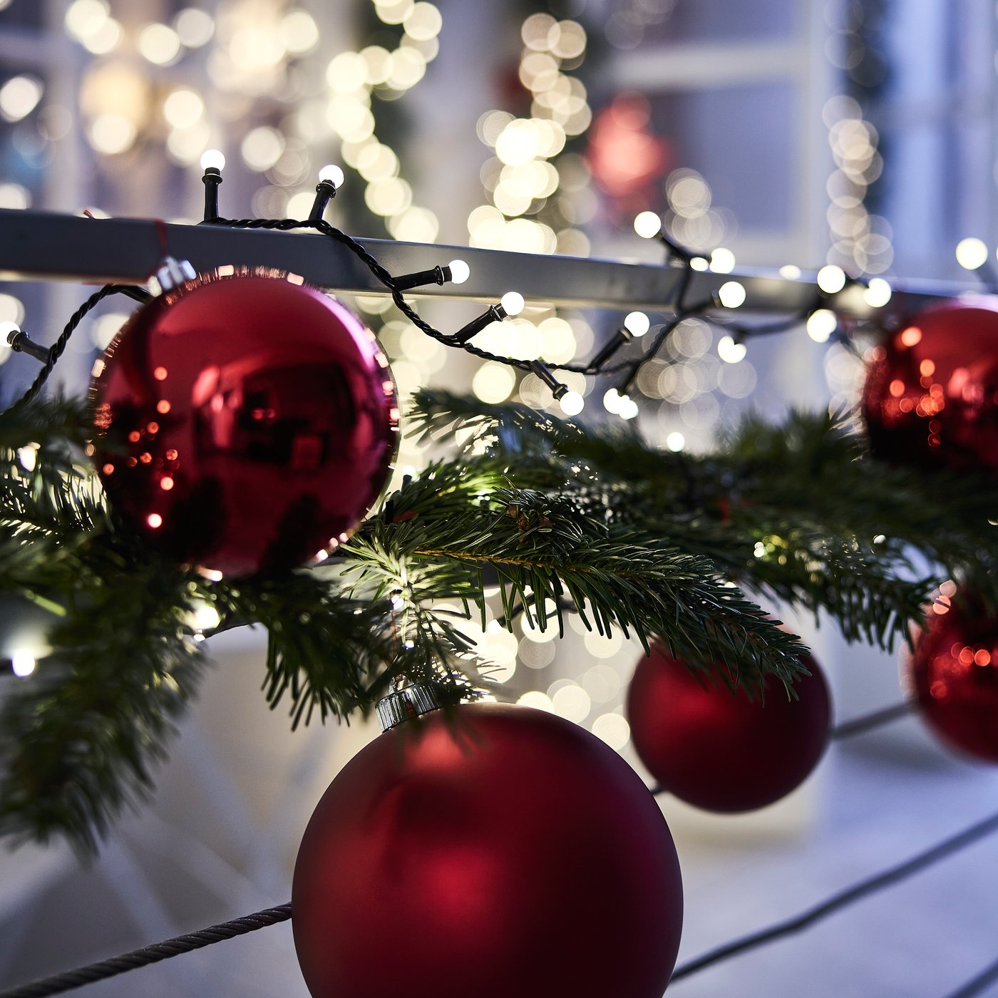 A lit outdoor STRÅLA lighting chain, a green VINTERFINT artificial garland and large red tree decorations on a balcony rail.