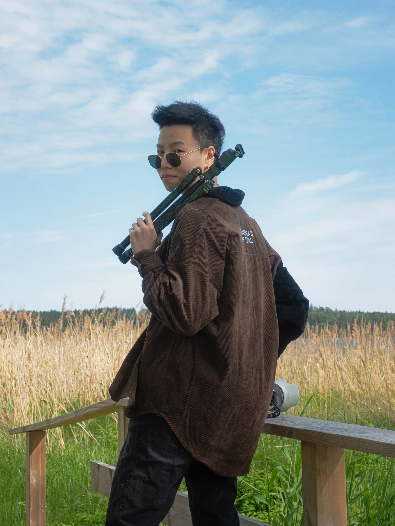 Žena s naočarima za sunce stoji pored polja obraslog travom i drži stativ za kameru na ramenu.