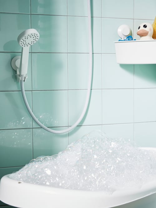 Dans une douche en carrelage, gros plan d’une douchette LILLREVET blanche permettant d’économiser l’eau, fixée à un support au-dessus d’une baignoire pour bébé.