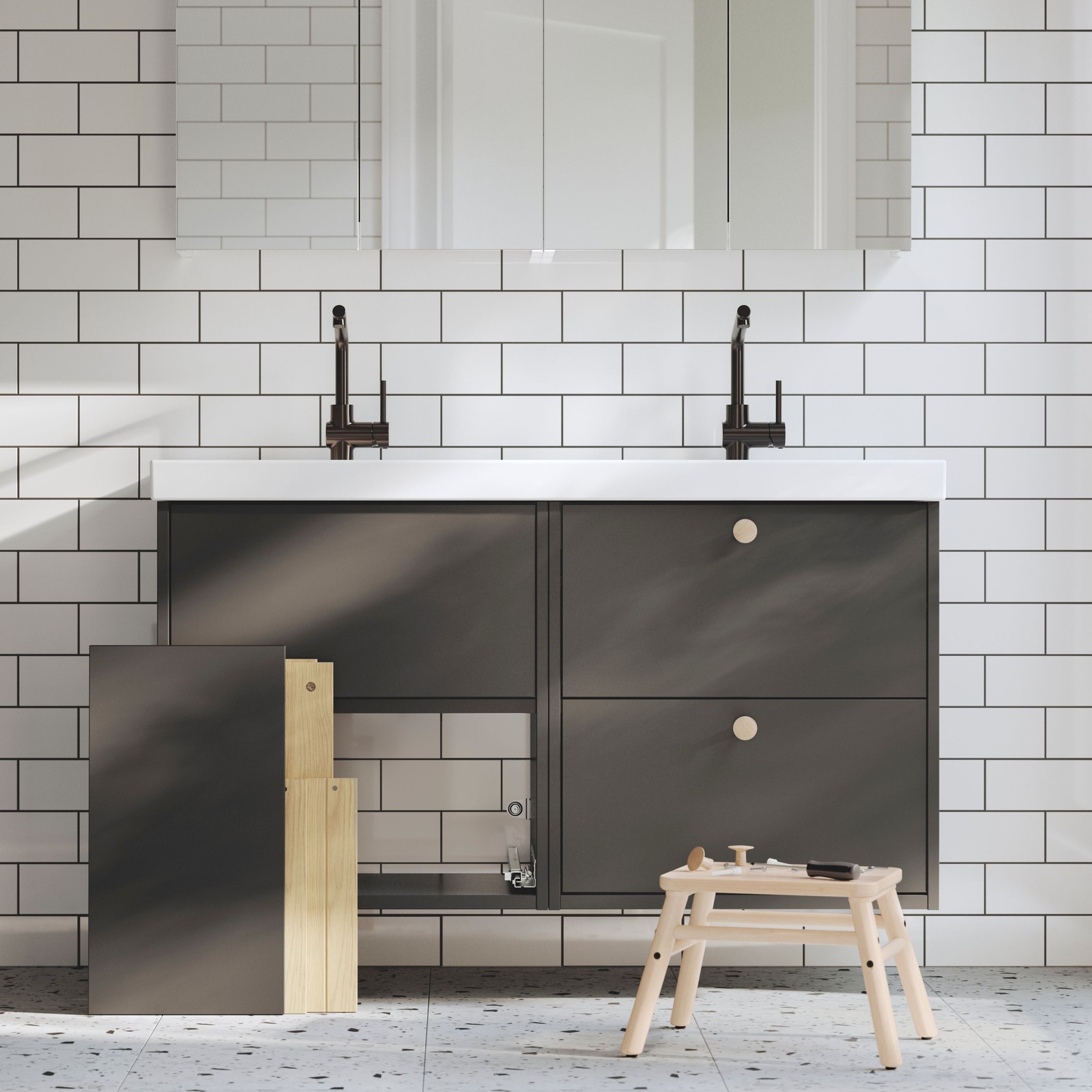 A bathroom has two dark grey HAVBÄCK wash-stands, one finished and one being assembled, and there are tools on a stool.