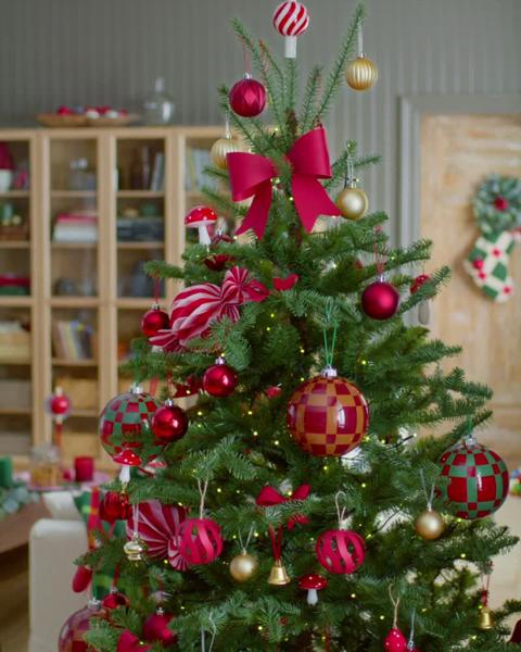 Un sapin de Noël décoré de boules rouges et dorées, de cannes en sucre et d’un grand nœud rouge est orné par une personne dans une pièce chaleureuse avec des étagères en bois remplies de livres et d’objets.