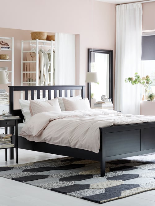 Cadre de lit HEMNES noir-brun avec une housse de couette rose, installé au milieu d’une chambre, à côté d’une table de chevet assortie.
