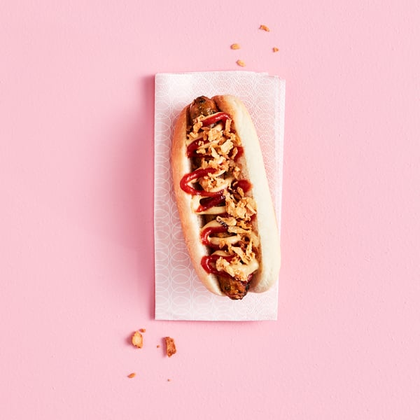 Ein KORVMOJ Gemüsewürstchen vegan mit ROSTAD LÖK Röstzwiebeln auf einem KORVBRÖD Hotdogbrötchen, serviert auf einer Papierserviette.