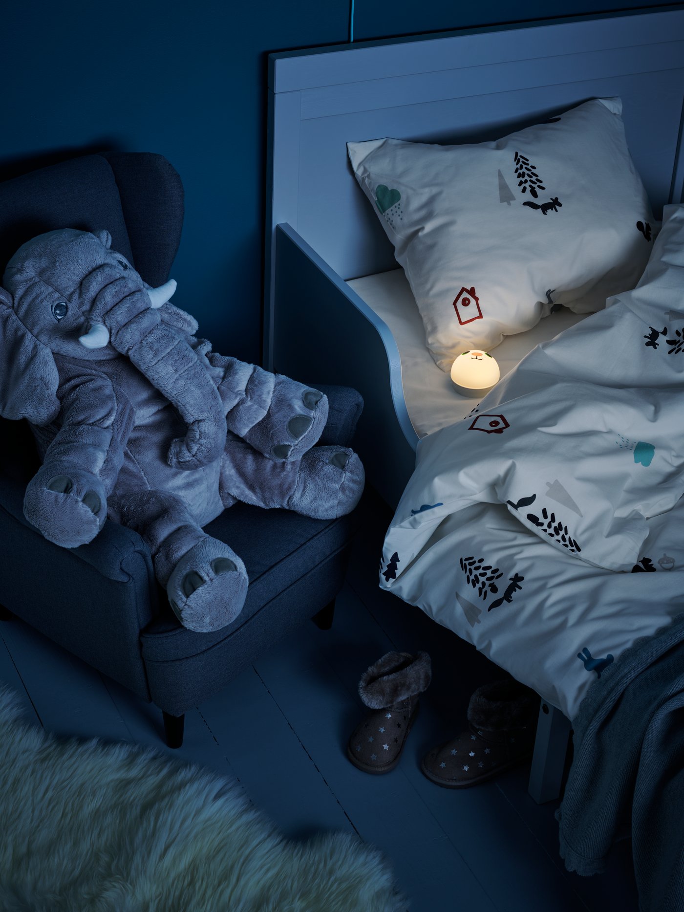 Un elefante de peluche JÄTTESTOR sentado en un sillón infantil junto a una cama con una luz de noche KORNSNÖ encendida.