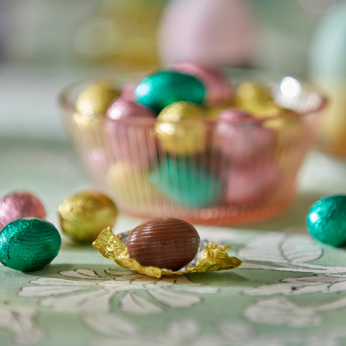 A close-up of a VÅRKÄNSLA milk chocolate egg, on top of a green, floral-patterned SMÖRFISK tablecloth.