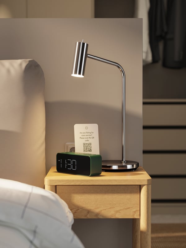 A hotel room with a TONSTAD bedside table in oak veneer with a VIRRMO work lamp and a green, digital BONDTOLVAN alarm clock.