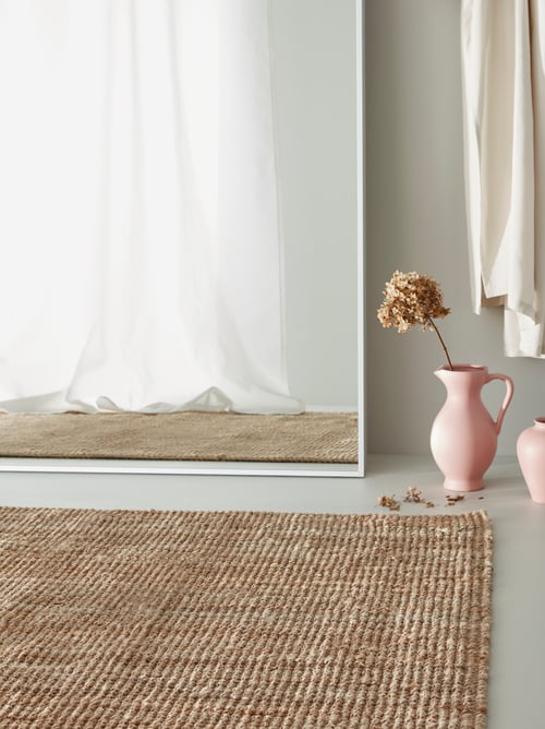 Wohnbereich mit einem LOHALS flach gewebten Teppich aus Naturjute, u. a. mit einer rosafarbenen Vase und einem großen Standspiegel, in dem sich lange Gardinen spiegeln.