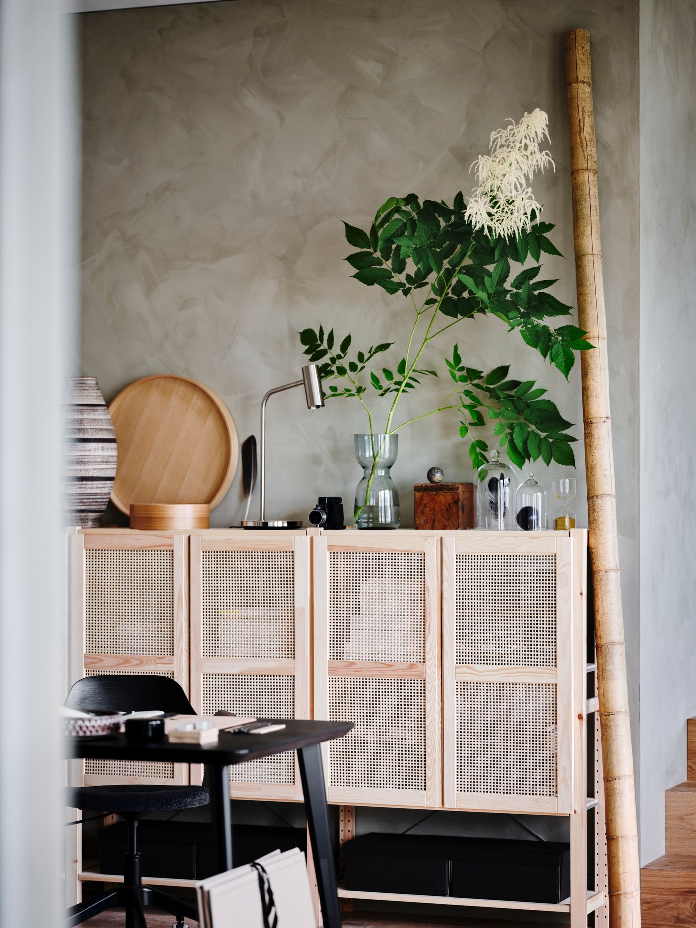 Espacio de trabajo con armarios con puertas beige, una lámpara de trabajo, una planta en un jarrón y una mesa rectangular.