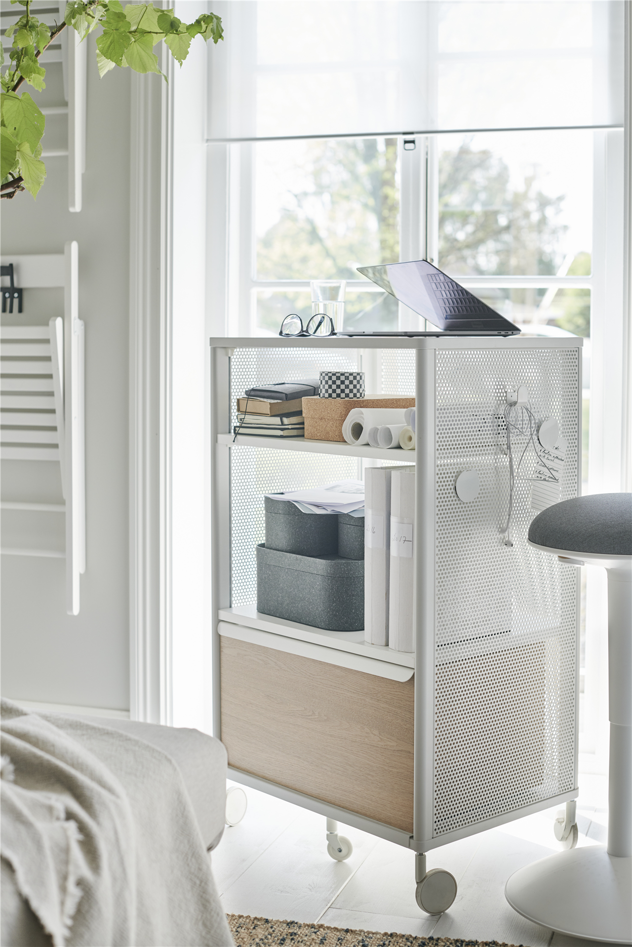 Ein BEKANT Aufbewahrungselement mit Rollen und Netz in Weiß neben einem Fenster. Auf dem Element ist ein Laptop zu sehen.