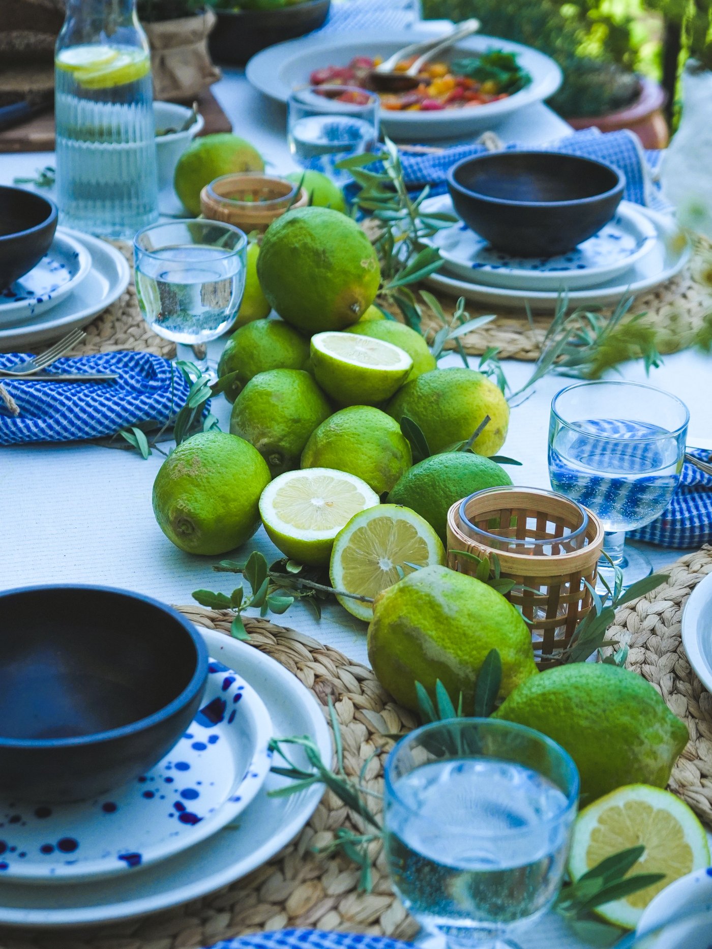 Une table dressée dans un style méditerranéen avec des assiettes IKEA Silvereda, des couverts NATTKLOT, des verres à pied FRUKTDUVA, des photophores PÅVEFINK, des citrons verts, des olives et une carafe à eau.