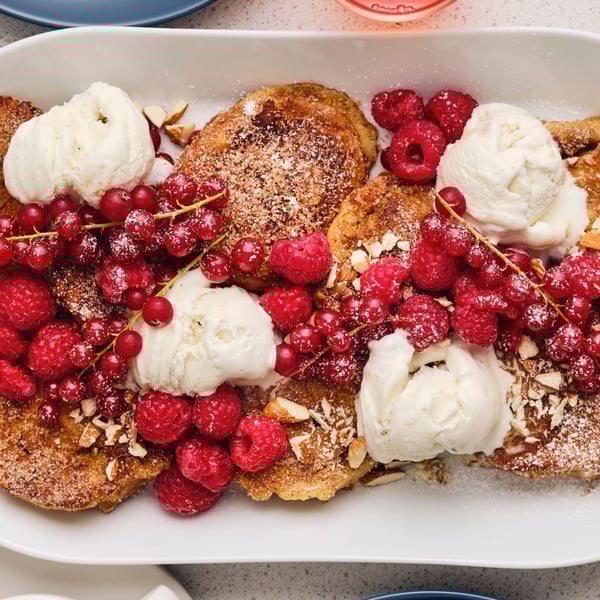 Auf einem weißen ovalen Teller liegen mehrere Stücke French Toasts mit Eis, frischen Himbeeren, roten Johannisbeeren und Mandeln darauf.