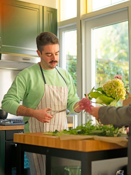A man wearing a HILDEGUN apron cutting flowers and preparing a floral arrangement in his kitchen.