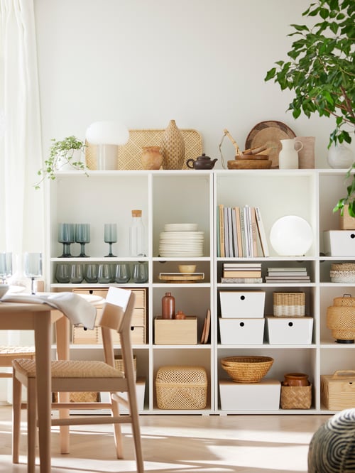 A dining room featuring a VIHALS shelving unit with 6 shelves, beige chairs, boxes with lids, wine glasses, and table lamps.