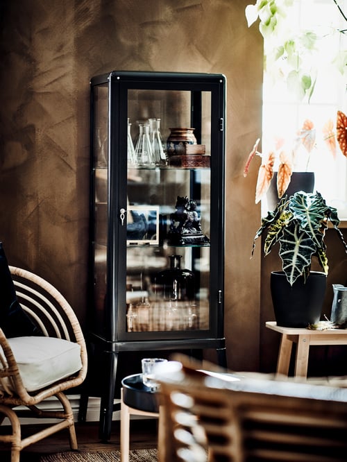 Salon avec une vitrine grise contenant des vases, un fauteuil beige, une plante en pot et une table basse ronde.