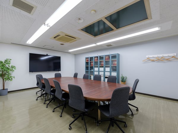 楕円形の木製テーブルを囲んでグレーのオフィスチェアを並べた会議室。壁に大型モニターと収納棚がある。