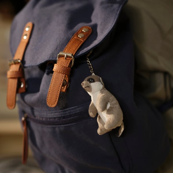 A beige SANDLÖPARE mini meerkat hangs as a mascot on a blue backpack with brown straps.