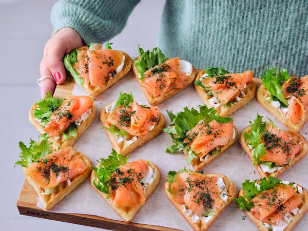 A person in a blue sweater is holding a wooden board which has twelve waffles with salmon, cream cheese and lettuce on it.