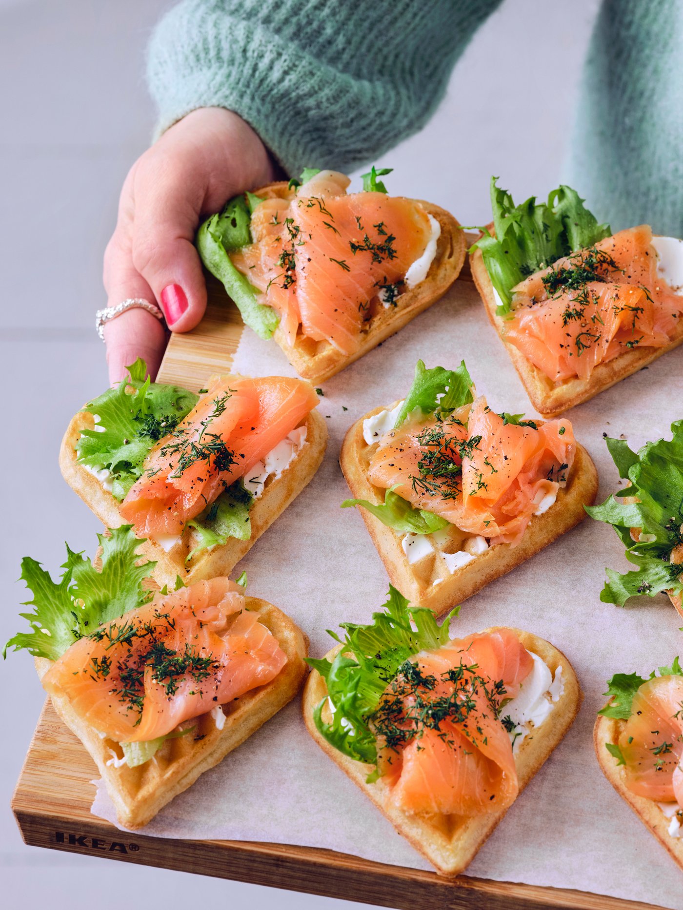 Eine Person in einem blauen Pullover hält ein Holzbrett in der Hand, auf dem zwölf Waffeln mit Lachs, Frischkäse und Kopfsalat liegen.