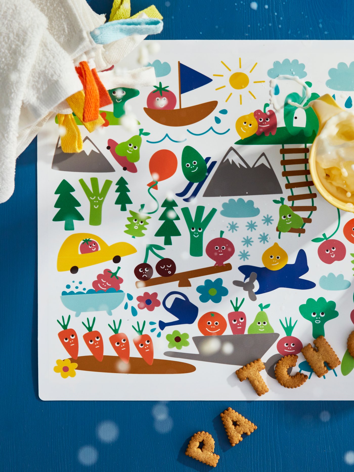 Milk is being splashed from a mug on a colourful MATVRÅ place mat on which some biscuits and KRAMA washcloths are lying.