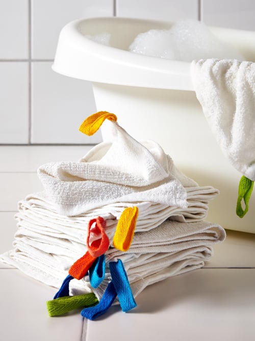 A pile of KRAMA washcloths in white with coloured tags beside a baby bath. A cloth is hanging on the side of the bath.