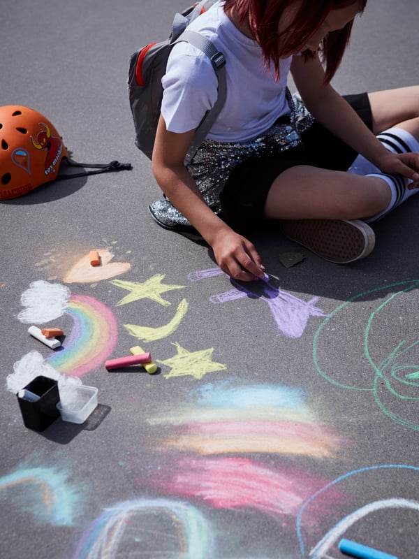 A young girl sitting with a backpack on the concrete ground drawing rainbows and shapes with coloured MÅLA chalks.