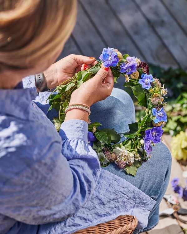 Eine Frau sitzt auf einem Stuhl und bindet einen Blumenkranz mit violetten Blüten