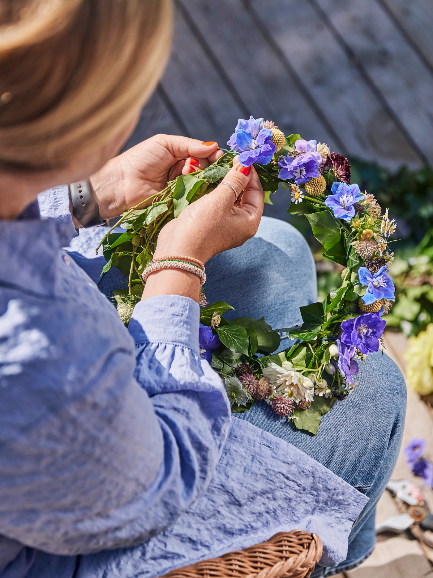 A person making a flower crown made of blue flowers and green leaves.