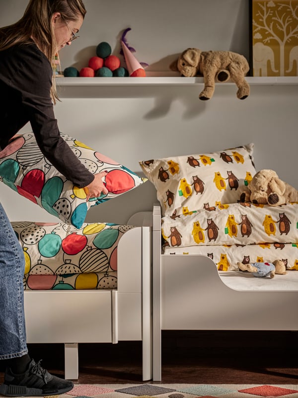 A woman making two white extendable beds with multicolour BRUMMIG and BUSENKEL bed linen, plus picture ledges on the wall.
